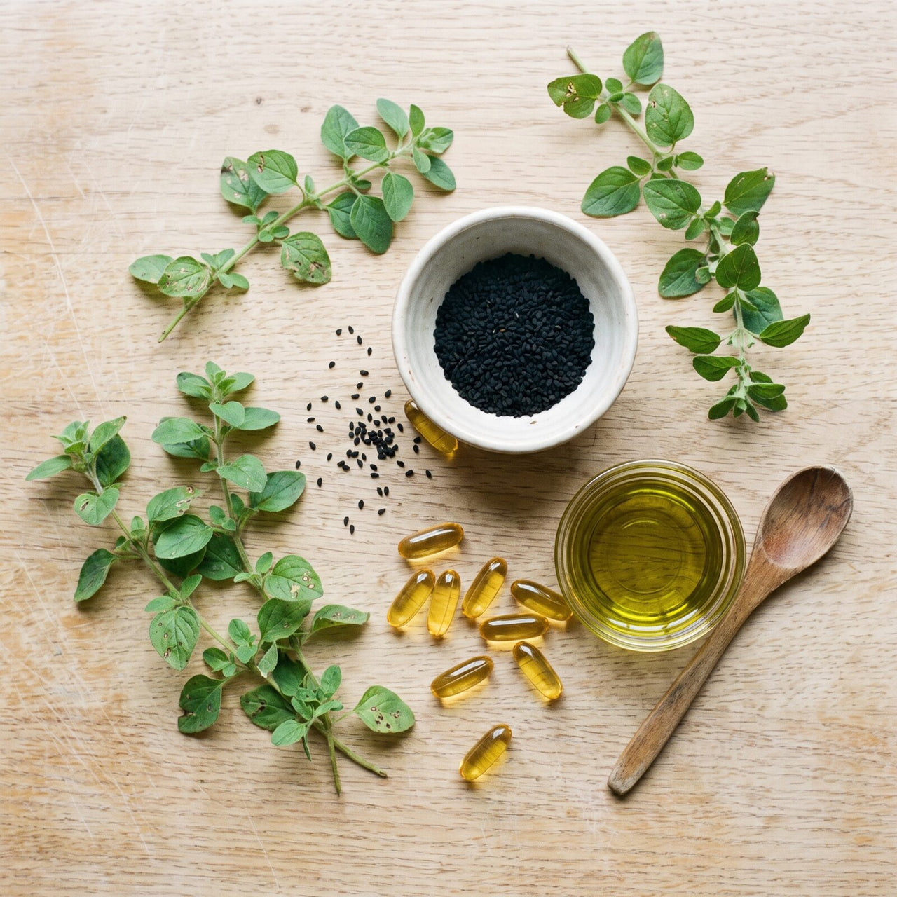 Fresh oregano, a bowl of black seed, golden oil and softgels on an oak table